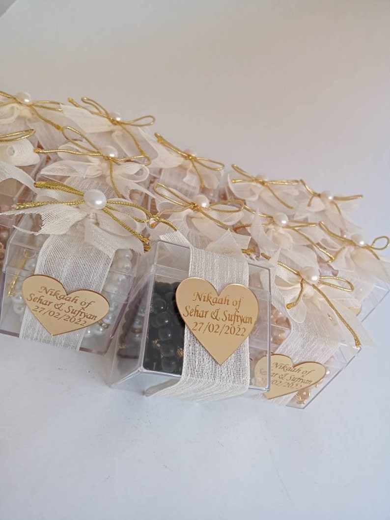May include: A group of small, clear boxes with white fabric and gold ribbon bows. Each box contains black beads and a gold heart-shaped tag with the text "Nikaah of Sehar & Sufiyan 27/02/2022".