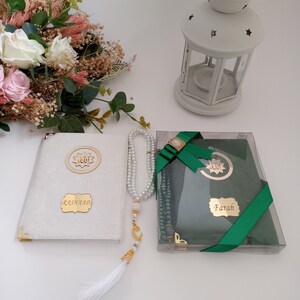 May include: A white velvet-covered book with gold corners and a gold plaque reading Rashaad, a string of white pearl prayer beads with a gold crescent charm and white tassel, and a green book in a clear box with a gold plaque reading Farah.