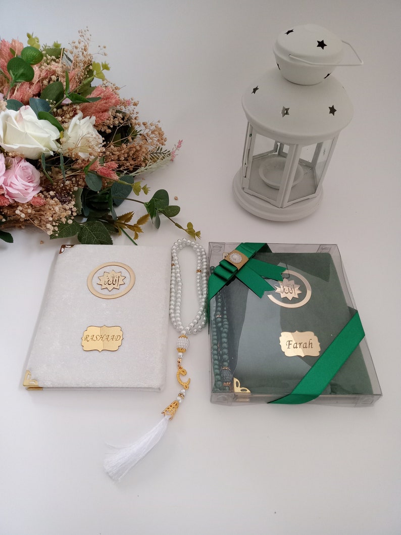 May include: A white velvet-covered book with gold corners and a gold plaque reading Rashaad, a string of white pearl prayer beads with a gold crescent charm and white tassel, and a green book in a clear box with a gold plaque reading Farah.