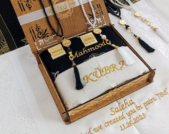 May include: A wooden box with a white and black prayer rug inside. The box is decorated with gold lettering and the words "Mahmood" and "Kübra". The rug has the words "Saleha" and "And we created you in pairs. 78:8" embroidered on it. The date "11.26.2023" is also embroidered on the rug.
