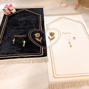 May include: Two black and white prayer rugs with gold trim and fringe. The rugs have a heart shape with a tulip design and the names "Mohamed" and "Ayesha" embroidered on them.