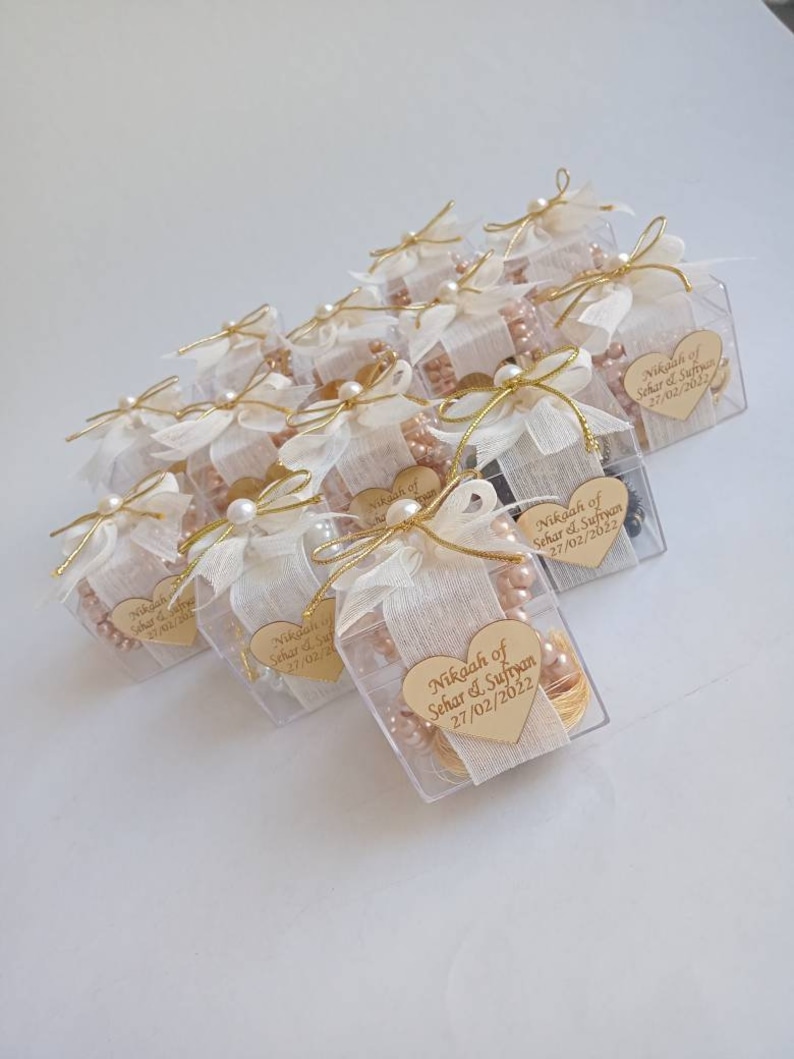 May include: A collection of small, clear boxes tied with white ribbon and gold string. Each box contains a small heart-shaped wooden tag with the text "Nikaah of Sehar & Sufiyan 27/02/2022".
