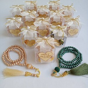May include: A set of wedding favors featuring small clear boxes tied with white ribbon and pearl accents. Each box contains a string of prayer beads with a gold tassel. The boxes are decorated with a heart-shaped tag that reads "Nikah of Sehar & Sufryan 27/02/2022".