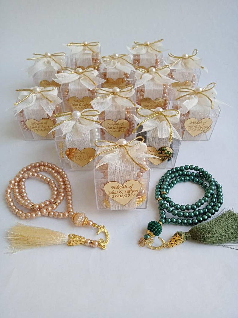 May include: A set of wedding favors featuring small clear boxes tied with white ribbon and pearl accents. Each box contains a string of prayer beads with a gold tassel. The boxes are decorated with a heart-shaped tag that reads "Nikah of Sehar & Sufryan 27/02/2022".