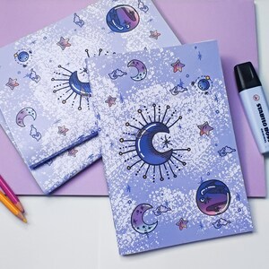 May include: Three purple notebooks with a white background and a moon and stars design. The notebooks are stacked on top of each other.