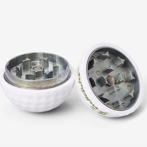 May include: Two white golf ball shaped grinders with a silver metal grinding mechanism. The grinders have a green logo that says "Swingers" on them.