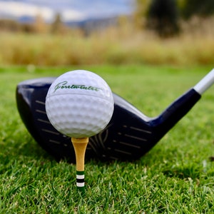 May include: A white golf ball with the text "Portwinter" printed on it, sitting on a tee and resting on a black golf club.