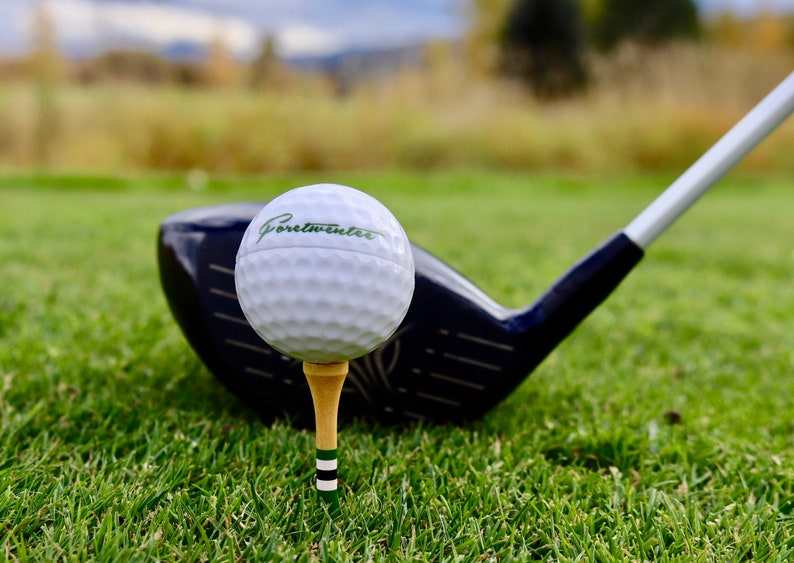 May include: A white golf ball with the text "Portwinter" printed on it, sitting on a tee and resting on a black golf club.