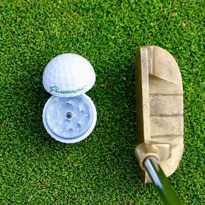 May include: A white golf ball with a green "Forewarning" logo on it, open to reveal a silver grinder inside. A gold putter sits on green grass.