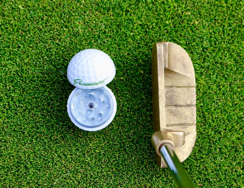 May include: A white golf ball with a green "Forewarning" logo on it, open to reveal a silver grinder inside. A gold putter sits on green grass.