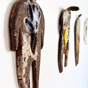 May include: A series of wall-mounted ceramic figures with textured, dark brown surfaces and cracked glaze details. The primary figure features a central blue accent, while others display yellow and multi-colored elements in a vertical orientation.