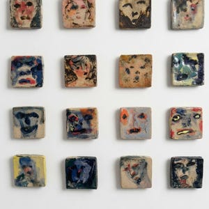 May include: A collection of sixteen square ceramic tiles arranged in a four-by-four grid. Each tile features a unique, hand-painted abstract face with varied colour palettes including shades of blue, red, yellow, black, and earthy tones on a light base.