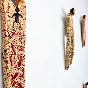 May include: Three elongated, wall-mounted ceramic figures with cream-coloured surfaces covered in black calligraphic script and red painted motifs. The figures feature dark hair, blue and yellow accents, and abstract human-like forms in a vertical arrangement.