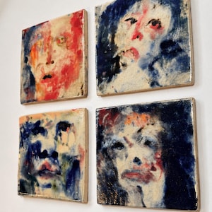 May include: A set of four square wall tiles featuring abstract portrait paintings. Each tile displays a human face rendered in expressive, blended brushstrokes of deep blue, vibrant red, and creamy white, creating a textured, layered appearance.