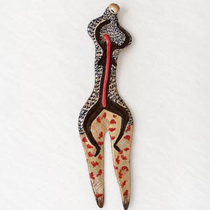 May include: A tall, slender, fork-like ceramic sculpture featuring a cream-coloured base with a black speckled pattern on the upper section. The lower portion displays small red leaf-like motifs, while a central vertical red line runs down the middle.