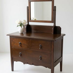 May include: A dark brown wooden vanity featuring a rectangular mirror supported by two vertical posts, two upper drawers, and one full-width lower drawer. The piece has decorative metal pulls and a scalloped apron. A white vase with white flowers sits on top.