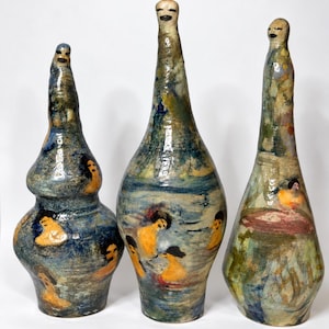 May include: Three tall, hand-formed ceramic vessels with elongated necks and rounded heads. The surfaces feature a mottled glaze in shades of blue, green, and yellow, decorated with painted figures of people swimming in water with dark hair.