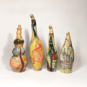 Może przedstawiać: Cztery ręcznie malowane rzeźby ceramiczne o różnych wysokościach i kształtach. Każdy element ma wąską szyjkę zwieńczoną małą, prostą twarzą. Powierzchnie ozdobione są kolorowymi abstrakcyjnymi wzorami, postaciami ludzkimi i rysunkami liniowymi w odcieniach żółci, błękitu, czerwieni i czerni.
