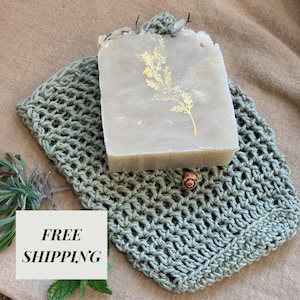 May include: A light-colored bar of soap with a gold floral design rests on a sage green crocheted washcloth. The soap is rectangular and has a textured surface. The washcloth is made of a loosely woven material.