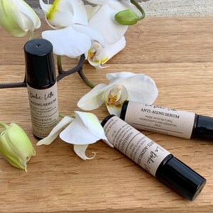 Puede incluir: Dos botellas de vidrio marr&oacute;n con tapones negros y etiquetas que dicen "La Dolce Villa Anti-Aging Serum" y "Hecho con ingredientes naturales y aceites esenciales". Las botellas est&aacute;n sobre una superficie de madera con flores de orqu&iacute;dea blancas.