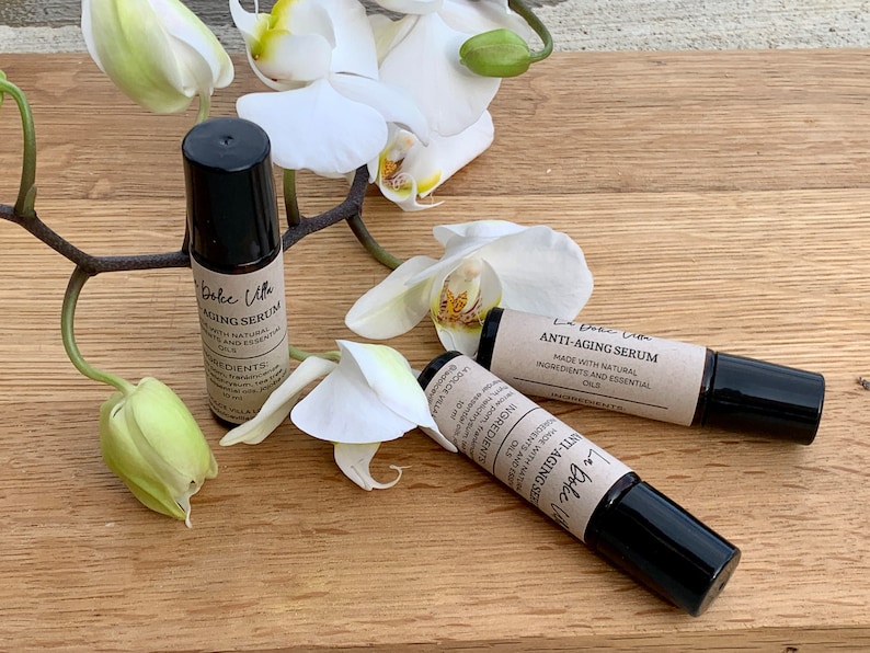 Puede incluir: Dos botellas de vidrio marr&oacute;n con tapones negros y etiquetas que dicen "La Dolce Villa Anti-Aging Serum" y "Hecho con ingredientes naturales y aceites esenciales". Las botellas est&aacute;n sobre una superficie de madera con flores de orqu&iacute;dea blancas.