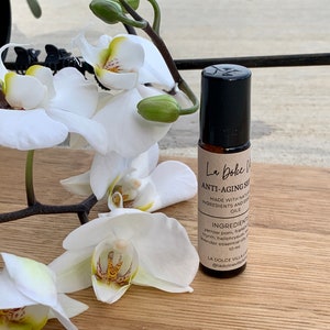 Puede incluir: Un frasco de vidrio marr&oacute;n con un aplicador de rodillo y una tapa negra. La etiqueta dice "La Dolce Villa Anti-Aging Serum" y enumera los ingredientes como "Yarrow, Pom, Incienso, Mirra, Helichrysum, Aceites esenciales de lavanda". El frasco est&aacute; sobre una superficie de madera con una orqu&iacute;dea blanca en el fondo.