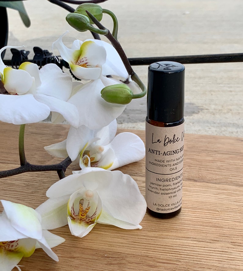 Puede incluir: Un frasco de vidrio marr&oacute;n con un aplicador de rodillo y una tapa negra. La etiqueta dice "La Dolce Villa Anti-Aging Serum" y enumera los ingredientes como "Yarrow, Pom, Incienso, Mirra, Helichrysum, Aceites esenciales de lavanda". El frasco est&aacute; sobre una superficie de madera con una orqu&iacute;dea blanca en el fondo.