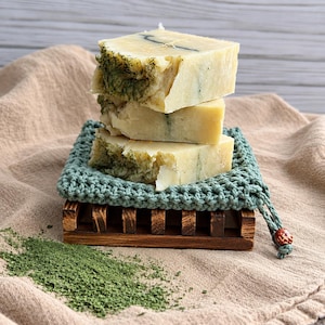 May include: A stack of three rectangular bars of off-white soap with visible green herbal flecks, resting on a square sage green crocheted mat placed atop a dark brown wooden slatted soap dish, with loose green herbs scattered nearby.