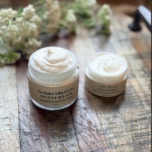 May include: Two glass jars of La Dolce Villa Living mineral sunscreen cream with white, whipped-texture product visible. The larger jar features a tan label with black text reading La Dolce Villa Living SUNSCREAM mineral sunscreen cream.