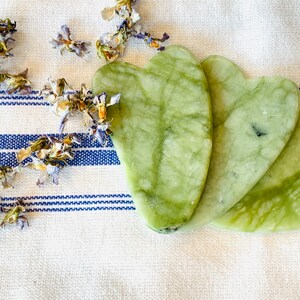 Puede incluir: Tres herramientas de gua sha en forma de coraz&oacute;n de jade verde sobre un fondo de tela a rayas blancas y azules.
