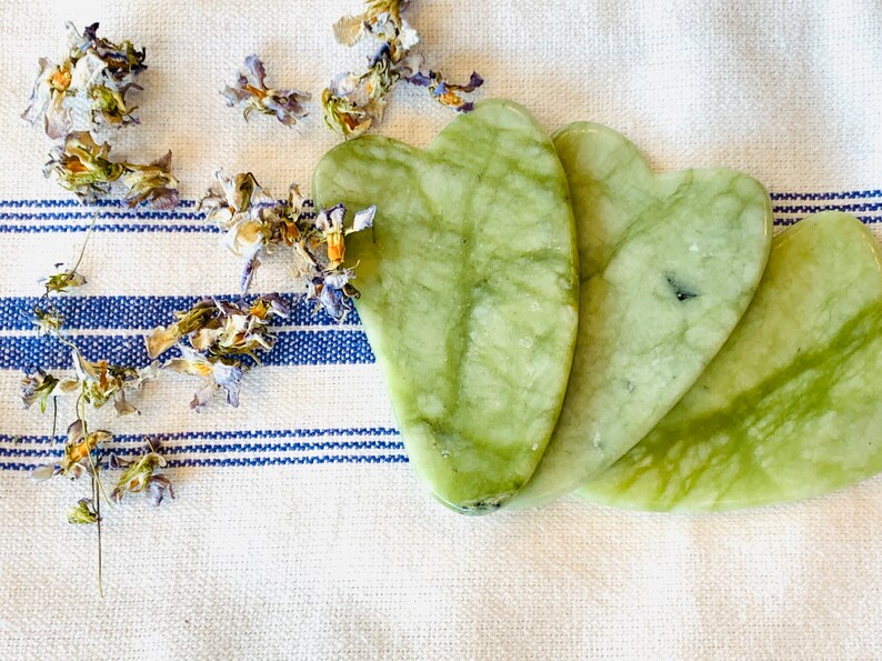 Puede incluir: Tres herramientas de gua sha en forma de coraz&oacute;n de jade verde sobre un fondo de tela a rayas blancas y azules.