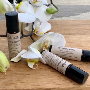 Puede incluir: Dos botellas de vidrio marr&oacute;n con tapones negros y etiquetas que dicen "Dolce Villa Anti-Aging Serum" y "Hecho con ingredientes naturales y aceites esenciales". Las botellas est&aacute;n sobre una superficie de madera con flores de orqu&iacute;dea blancas.