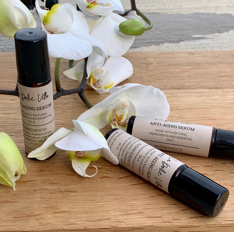 Puede incluir: Dos botellas de vidrio marr&oacute;n con tapones negros y etiquetas que dicen "Dolce Villa Anti-Aging Serum" y "Hecho con ingredientes naturales y aceites esenciales". Las botellas est&aacute;n sobre una superficie de madera con flores de orqu&iacute;dea blancas.