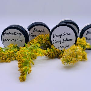 May include: Four small sample jars with black lids and white labels. The labels have black text that reads "Hydrating face cream", "Sample Sun", "Hemp Seed Body Lotion sample", and "Natural Deodorant sample". The jars are sitting on a white surface with yellow flowers in front of them.