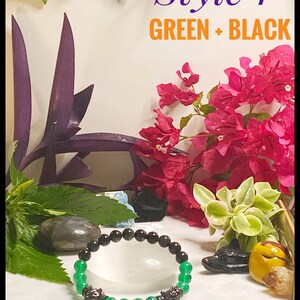 May include: A green jade and black obsidian beaded bracelet with wolf head charms. The bracelet is on a white surface with green and purple plants and flowers.