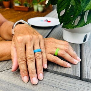 May include: Two hands with a light blue and a green silicone ring. The rings are on the ring fingers. A silver bracelet is on the left wrist. A white plate with small colorful objects is in the background.