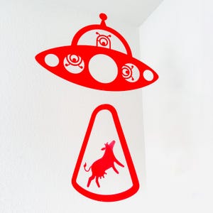 May include: Red metal UFO with three round windows and a small alien inside, hanging from the ceiling above a red metal cow silhouette.