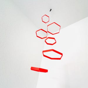 May include: A red geometric mobile with hexagonal shapes hanging from the ceiling. The mobile is made of paper or cardboard and is suspended by thin strings.