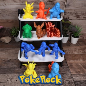 Può includere: Una mensola a più livelli espone figurine colorate stampate in 3D. Le figurine sono in vari colori, tra cui giallo, arancione, blu, verde e marrone. La mensola è bianca e nera e la base presenta il testo "PokeRock" in blu e giallo.