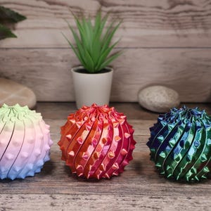 May include: Three decorative, spiky spheres. The left sphere has a green to pink to blue gradient. The middle sphere is red and orange. The right sphere is blue and green. They are displayed on a wooden surface.