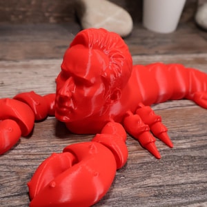 May include: A vibrant red sculpture of a lobster with a human face. The lobster features large claws and a segmented body. The sculpture is made of a solid material and rests on a wooden surface.