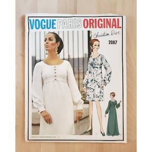May include: Vogue Paris Original sewing pattern by Christian Dior, pattern number 2087. The pattern features three illustrations of women wearing dresses. The first dress is white with gold buttons, the second dress is green with a floral print, and the third dress is a solid green.