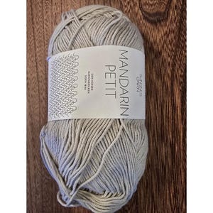 May include: A skein of light gray yarn with a white label. The label reads "MANDARIN PETIT" in large, bold letters. The yarn is tightly wound and appears soft. The label also includes the text "SANDNES GARN" and other text.