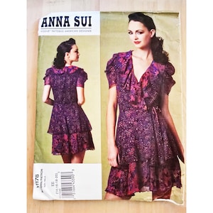 May include: A vintage Anna Sui Vogue pattern featuring a purple dress with a floral print. The dress has a ruffled neckline and sleeves, with a tiered skirt. The pattern includes two images of the dress and the text "ANNA SUI" and "VOGUE PATTERNS AMERICAN DESIGNER".