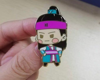 May include: A metal enamel pin featuring a stylized cartoon character with long black hair tied in a high ponytail, wearing a bright pink headband, a teal and white tunic with a pink sash, and pink circular blush marks on the cheeks.