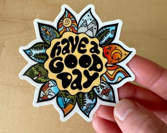May include: A white sticker with a yellow sun in the center. The sun has the words "have a good day" written in black. The sun is surrounded by a colorful flower design with various nature scenes.