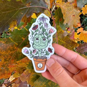 May include: A green cartoon plant with a face and flowers growing from it, in a brown pot with a peace sign. The plant is on a sticker.