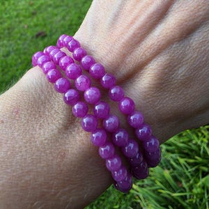 May include: A triple-strand stretch bracelet composed of small, round, polished magenta-colored beads. The beads feature a translucent quality with subtle internal variations in color and texture, arranged in three continuous loops around the wrist.