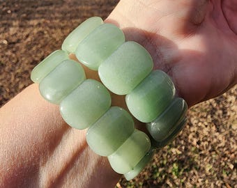 May include: A light green bracelet composed of large, rounded, rectangular beads. The beads have a smooth, polished appearance and are strung together to form a flexible band. The bracelet is shown on a wrist, with a natural outdoor background.
