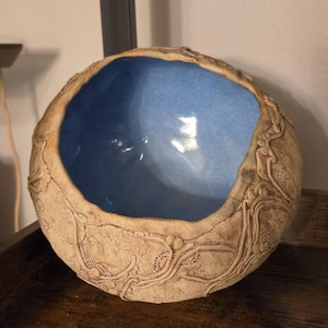 May include: A round ceramic bowl with a textured, tan-colored exterior featuring raised, intricate vine-like patterns. The interior of the bowl is finished with a smooth, glossy, light blue glaze that contrasts with the rough, earthy outer surface.